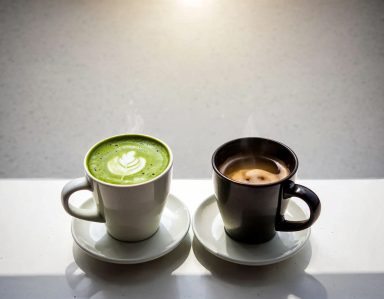 Zwei Tassen mit Heißgetränken stehen vertraut nebeneinander auf hellem Untergrund. Matcha Latte Art trifft schwarzen Kaffee.